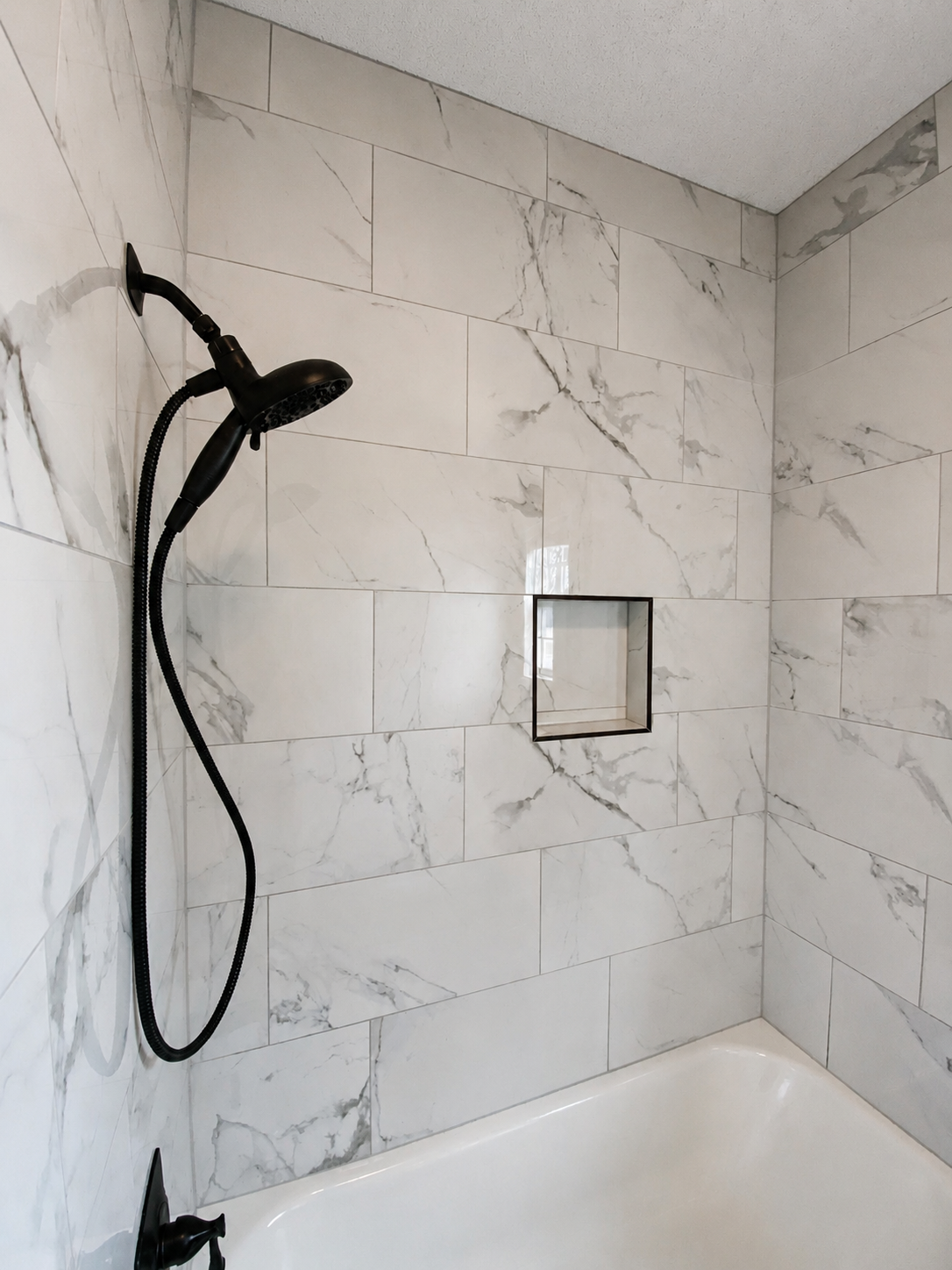 Completed tub and shower surround with marble-look tile, recessed niche, and matte-black fixtures