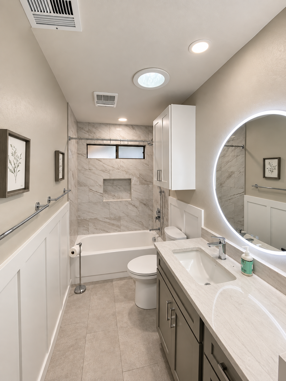 Completed bathroom remodel with a tiled tub and shower surround, gray vanity, illuminated round mirror, and white board-and-batten wainscoting